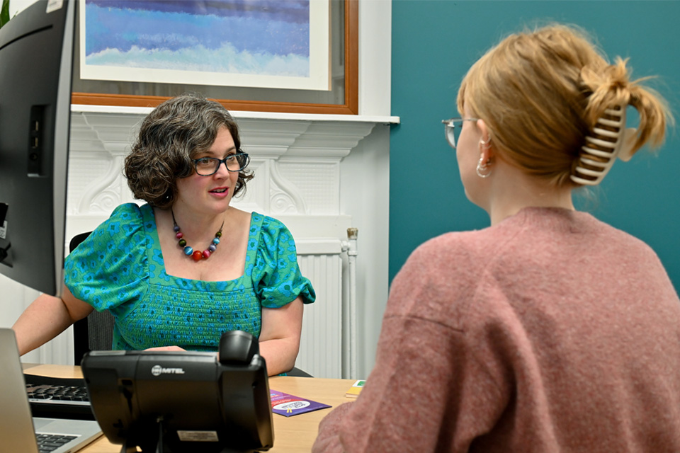 A women wearing a blue dress, listening to a student in a pink jumper, listening to student.